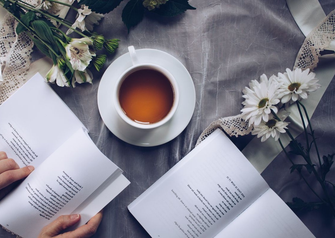 Top-down view of hands holding an open book beside a cup of tea, white alstroemeria and daisies on grey linen with lace ribbons