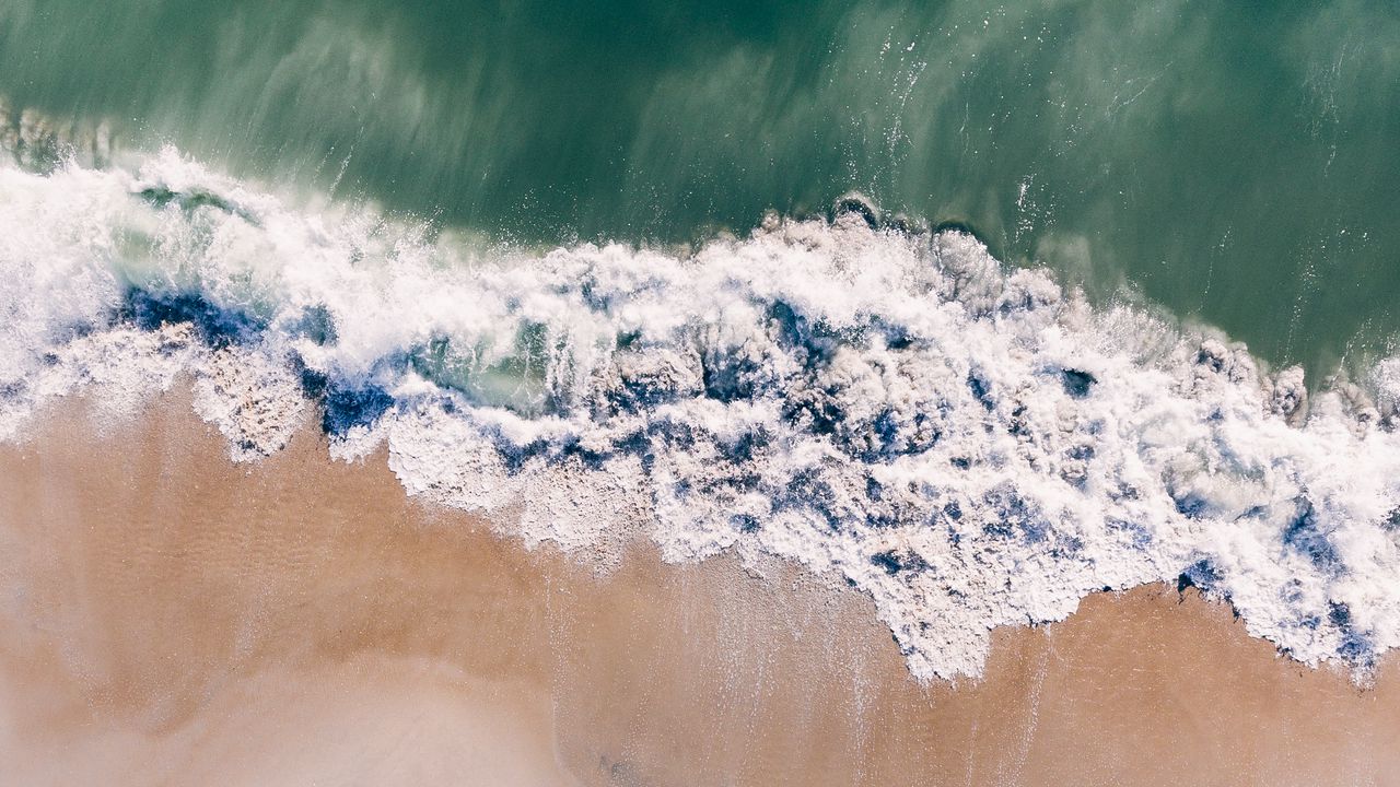 Top-down aerial photograph of teal ocean water, white seafoam waves, and light tan sand along a shoreline